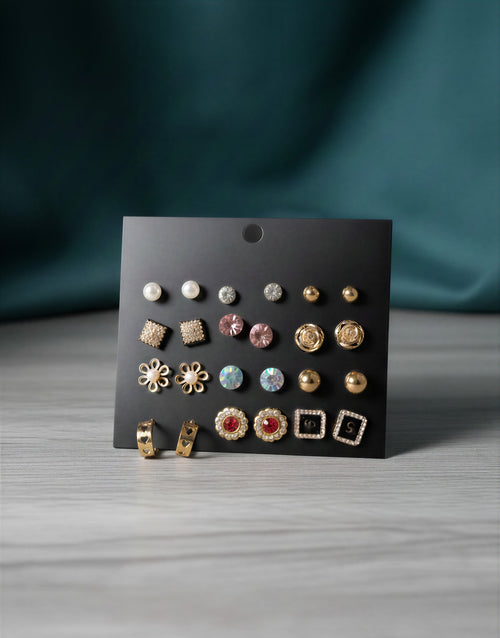 12 pairs of gold-tone earrings displayed on a black card including white pearls, pink crystals, floral studs, and heart-shaped gold hoops.