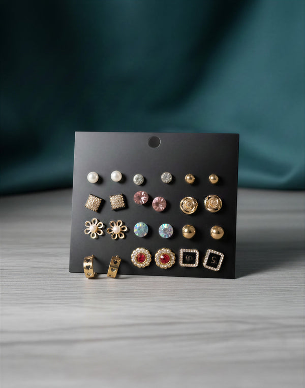 12 pairs of gold-tone earrings displayed on a black card including white pearls, pink crystals, floral studs, and heart-shaped gold hoops.